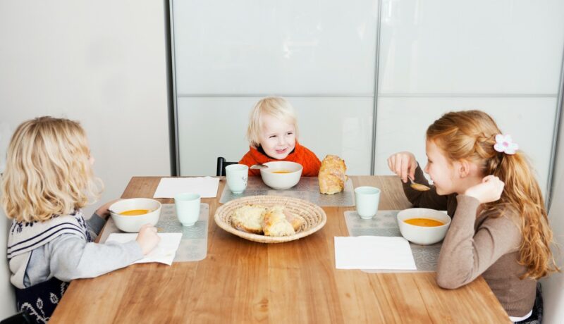 Trei copii zâmbitori stau la masa și servesc împreună prânzul