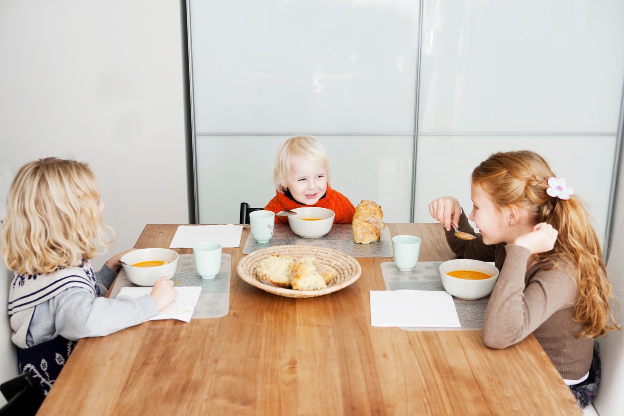 Trei copii zâmbitori stau la masa și servesc împreună prânzul