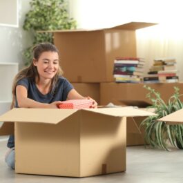 O femeie zâmbește în timp ce despachetează o cutie mare de carton, după ce s-a mutat singură