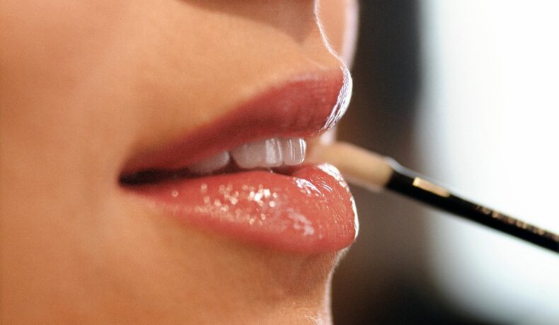 Cadru apropiat cu buzele unei femei, care aplică luciu de buze cu pensula