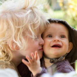 Un copil blond cu plete sărută pe obraz un bebelus zâmbitor cu pălărie