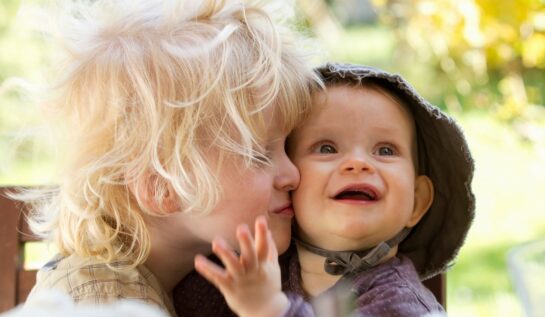 Un copil blond cu plete sărută pe obraz un bebelus zâmbitor cu pălărie