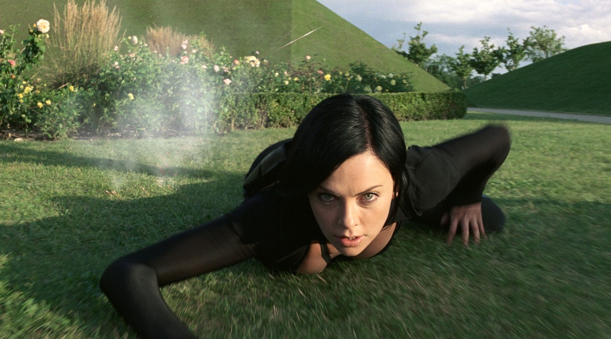 Charlize Theron stă întinsă pe iarbă. Are părul negru scurt și poartă un costum negru