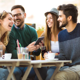 Un grup de patru prieteni care beau cafea împreună și râd, iar unul dintre ei ține în mână o tabletă
