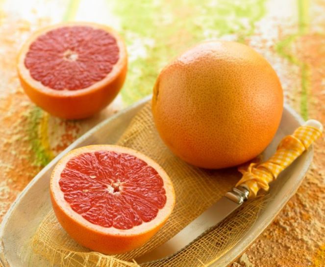 Două grapefruit roșii, așezate pe o tavă, lângă un cuțit, pe o suprafață colorată în mai multe nuanțe de portocaliu și verde