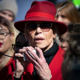 Jane Fonda participă la un protest îmbrăcată cu un palton roșu, cu o pălărie roție pe ca și cu ochelari de soare