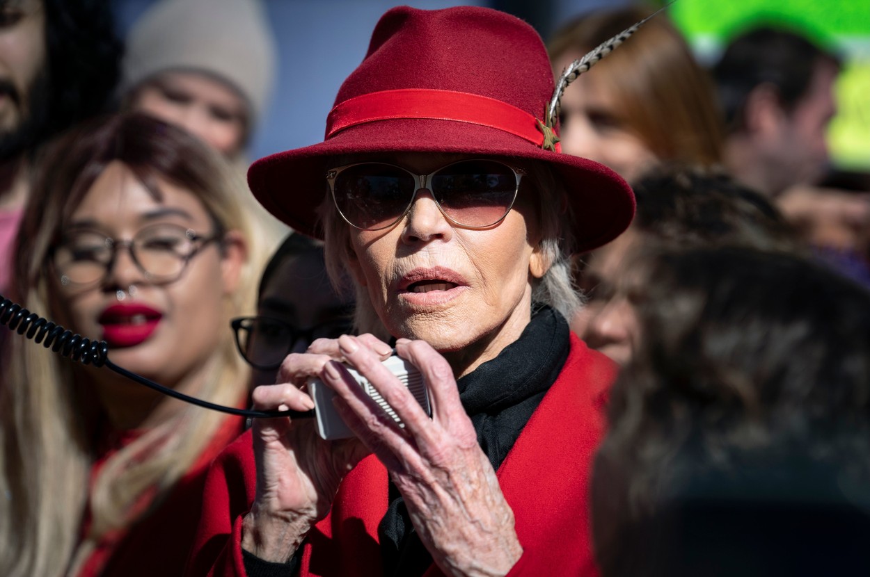 Jane Fonda participă la un protest îmbrăcată cu un palton roșu, cu o pălărie roție pe ca și cu ochelari de soare