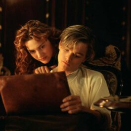 Leonardo Di Caprio îmbrăcat înr-o cămașă albă schițează un desen pe o planșă în timp ce Kate Winslet îl privește cu capul așezat pe umărul lui.
