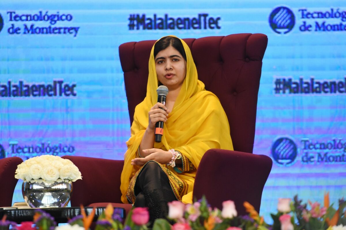Malala este îmbrăcată într-un sari galben și stă așezată pe un fotoliu roșu în timpul unei conferințe