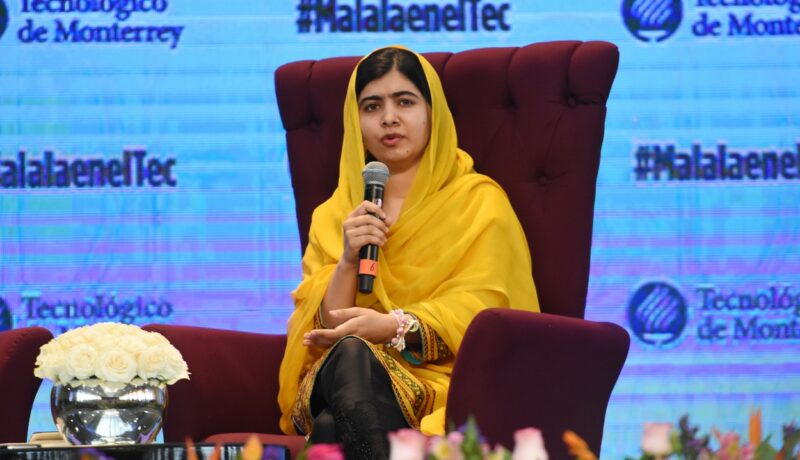 Malala este îmbrăcată într-un sari galben și stă așezată pe un fotoliu roșu în timpul unei conferințe