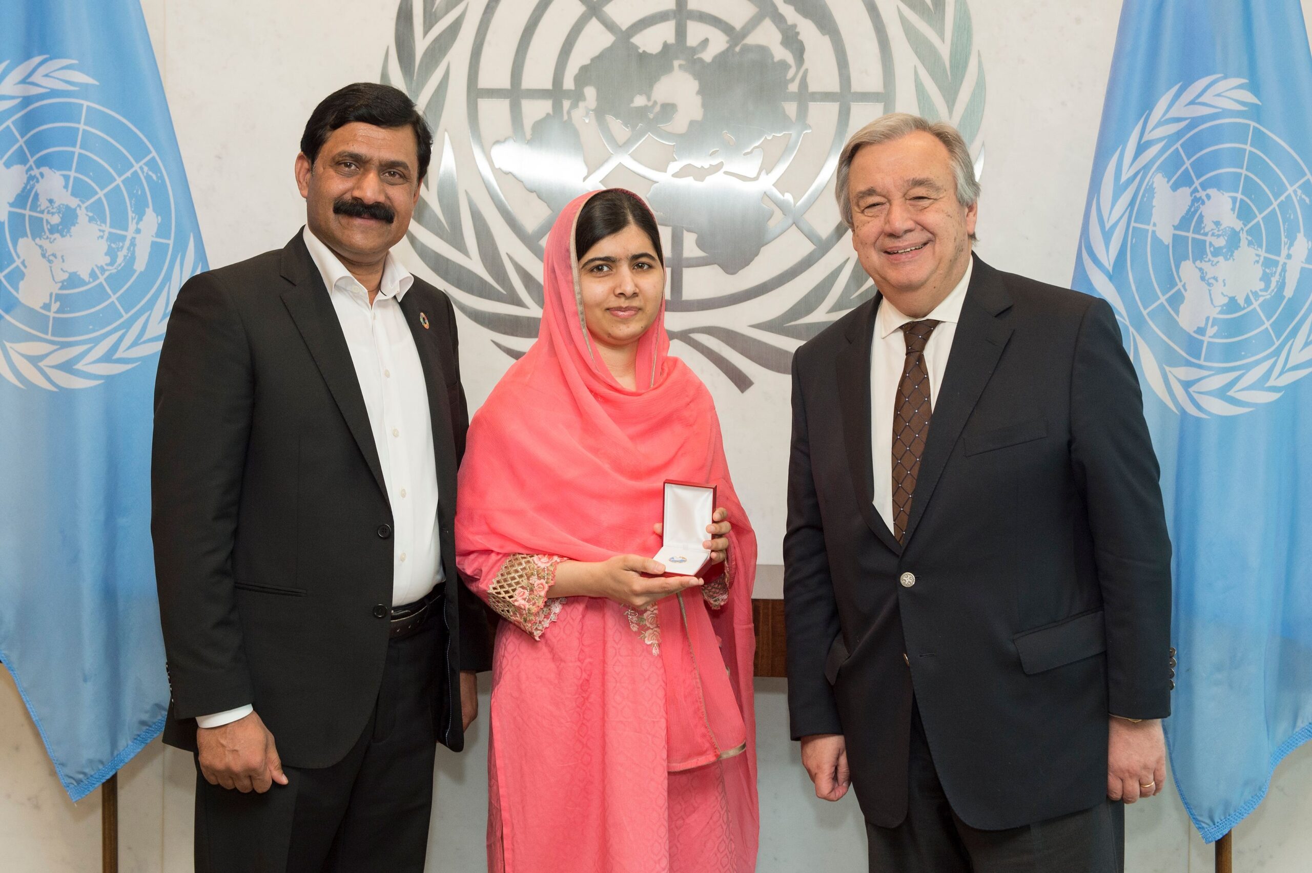 Malala pozează alături de tatăl ei și de secretarul general ONU la sediul organizației. Malala este îmbrăcată cu un sari roz și ce doi bărbați poartă costume negre.