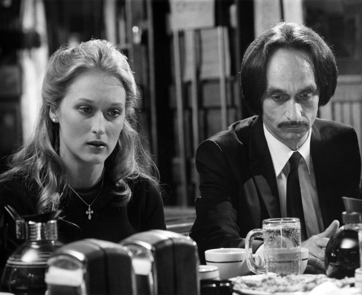 Fotografie alb-negru în care Meryl Streep stă așezată la masă alături de John Cazale