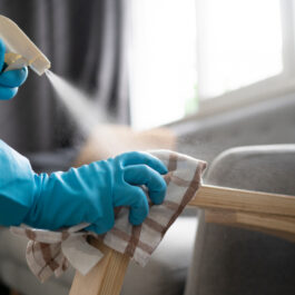Două mâini care poartă mănuși albastre pentru curățenie șterg cu o cârpă și un produs de curățare mânerul unui fotoliu