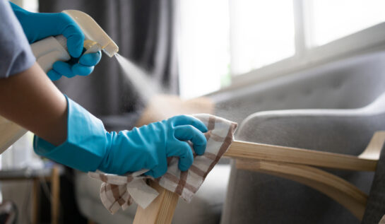 Două mâini care poartă mănuși albastre pentru curățenie șterg cu o cârpă și un produs de curățare mânerul unui fotoliu