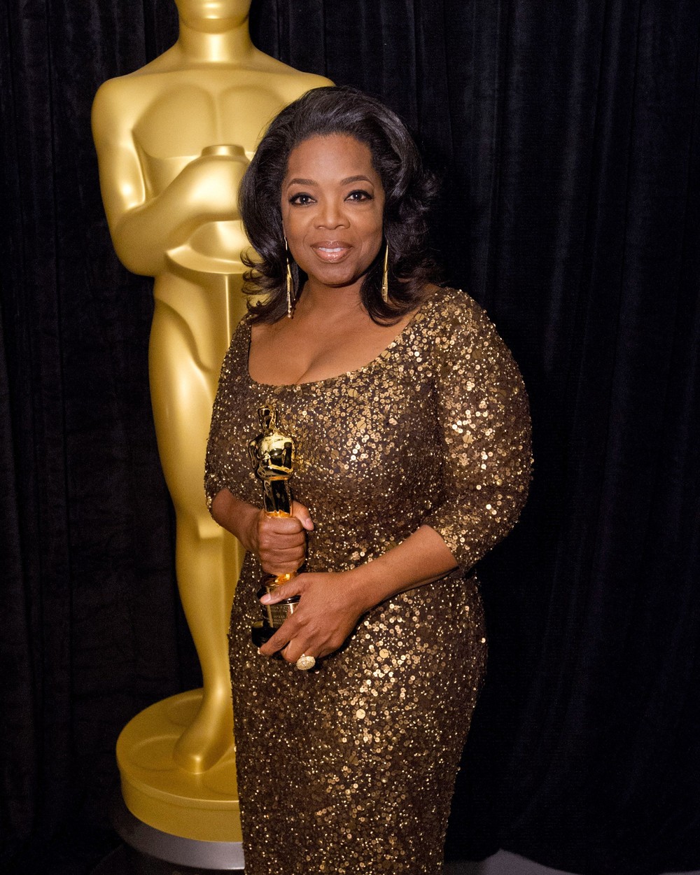 Oprah Winfrey este îmbrăcată într-o rochie aurie și ține în mână un trofeu Oscar