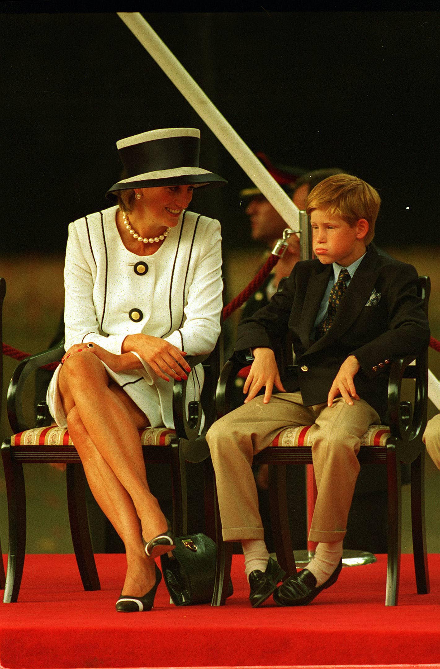 Prințesa Diana îmbrăcată ntr-un compleu alb elegant alcătuit din sacou și fustă participă la un eveniment alături de Prințul Harry îmbrăcat în pantaloni crem și sacou albastru