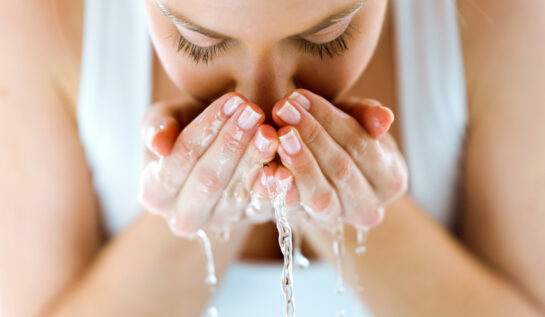 O femeie cu maiou alb, se spală pe față cu apă