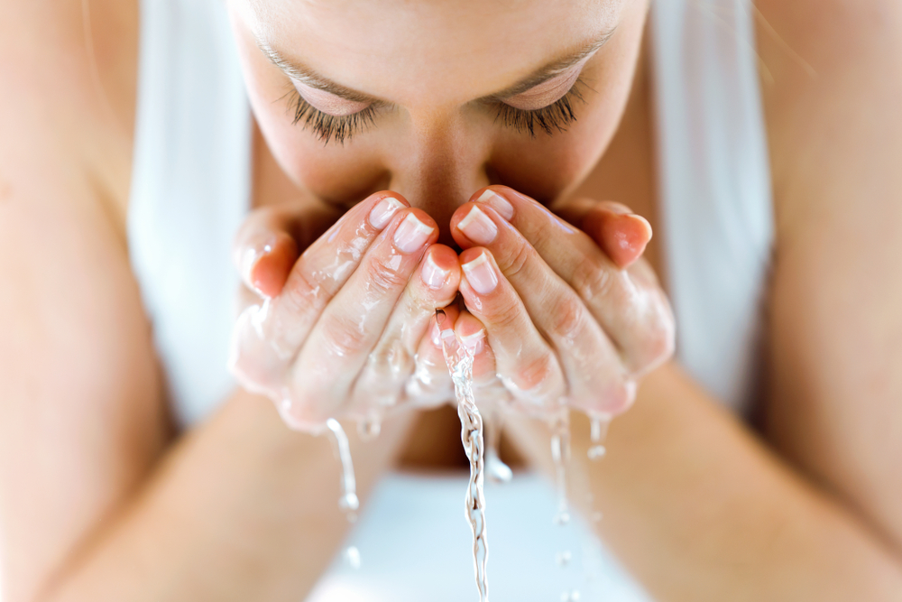 O femeie cu maiou alb, se spală pe față cu apă