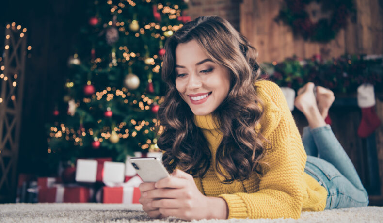 Femeie cu pulover galben, așezată lângă brad, ține telefonul în mână și zâmbește citind mesaje de Crăciun