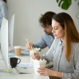 O femeie care mănâncă repede în timp ce stră la birou, în fața unui calculator.