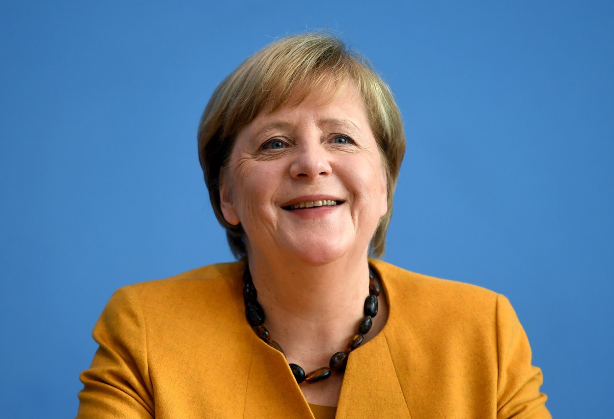 Angela Merkel zâmbește larg îmbrăcată într-un sacou galben și cu mărgele albastre la gât