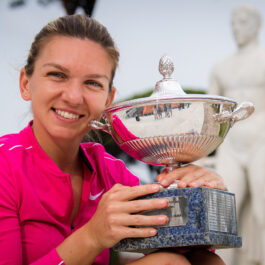 Simona Halep zâmbește și ține în mână un trofeu argintiu. Este îmbrăcată într-o bluză sport roz