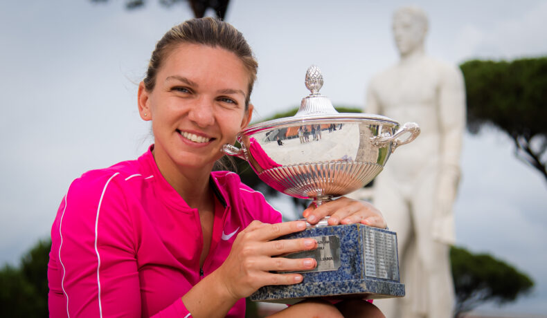 Simona Halep zâmbește și ține în mână un trofeu argintiu. Este îmbrăcată într-o bluză sport roz