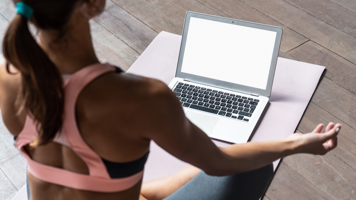 O femeie îmbrăcată cu o bustieră sport și colanți stă într-o poziție de yoga în fața unui laptop
