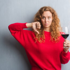 O femeie cu părul creț și blond îmbrăcată într-un pulover roșu ține în mână un pahar de vin roșu, iar cu cealaltă mână arată semnul dislike