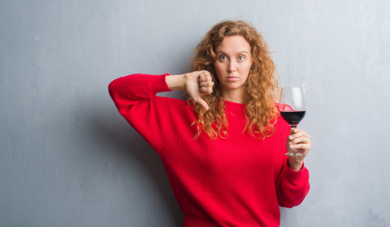 O femeie cu părul creț și blond îmbrăcată într-un pulover roșu ține în mână un pahar de vin roșu, iar cu cealaltă mână arată semnul dislike