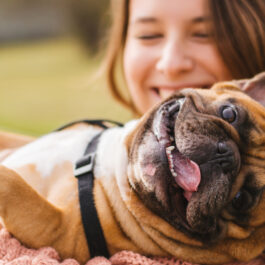 O femeie care zâmbește larg ține în brațe un câine maro care scoate limba afară