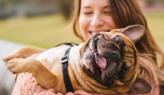 O femeie care zâmbește larg ține în brațe un câine maro care scoate limba afară