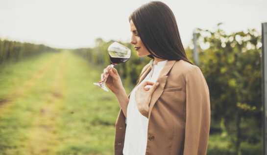 O femeie cu părul lung șaten bea un pahar de vin roșu pe o plantație de struguri