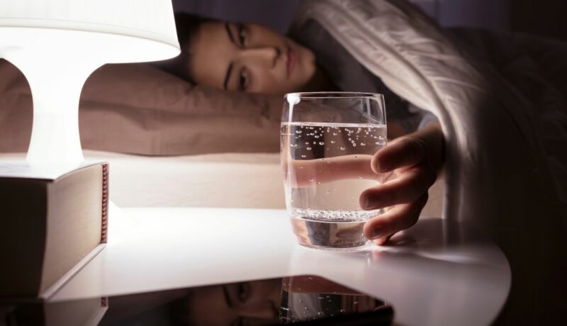 Femeie care stă noaptea în pat și ia un pahar cu apă de pe noptieră. Veioza luminează paharul cu apă și o carte.