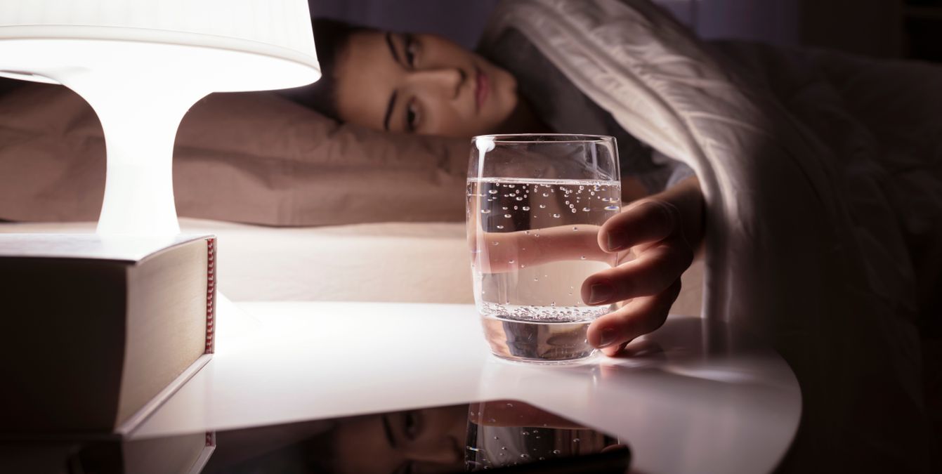 Femeie care stă noaptea în pat și ia un pahar cu apă de pe noptieră. Veioza luminează paharul cu apă și o carte.