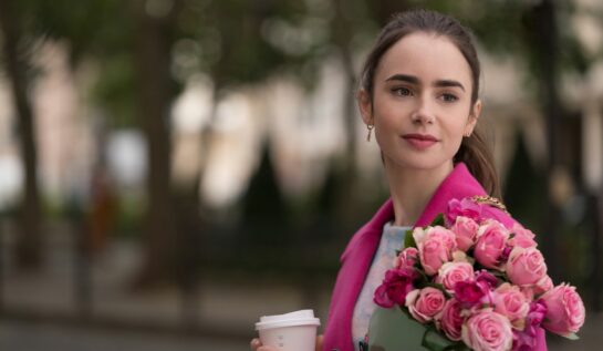 Lilly Collins în timpul filmărilor pentru Emily in Paris îmbrăcată cu un sacou roz și ține în mână un buchet de trandafiri roz
