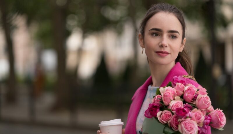 Lilly Collins în timpul filmărilor pentru Emily in Paris îmbrăcată cu un sacou roz și ține în mână un buchet de trandafiri roz