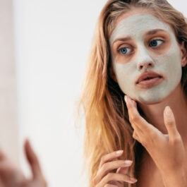 O femeie tânără cu părul lung și blond se uită în oglindă după ce și-a aplicat pe față o mască