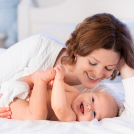 O mamă zâmbește în timp ce se uită la copilul ei care stă întins pe pat