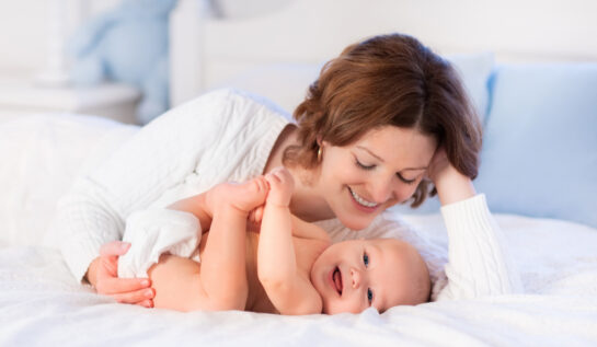 O mamă zâmbește în timp ce se uită la copilul ei care stă întins pe pat