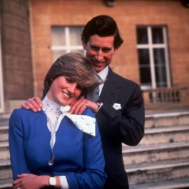 Prințul Charles îmbrăcat într-un costum negru și cravată cu buline o ține de umeri de la spate pe Prințesa Diana îmbrăcată cu un sacou albastru