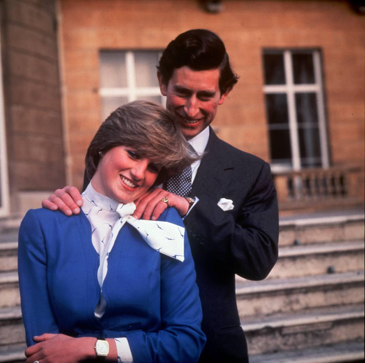 Prințul Charles îmbrăcat într-un costum negru și cravată cu buline o ține de umeri de la spate pe Prințesa Diana îmbrăcată cu un sacou albastru