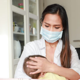 O femeie cu părul lung și șaten poartă o mască de protecție pe față și își alăptează copilul