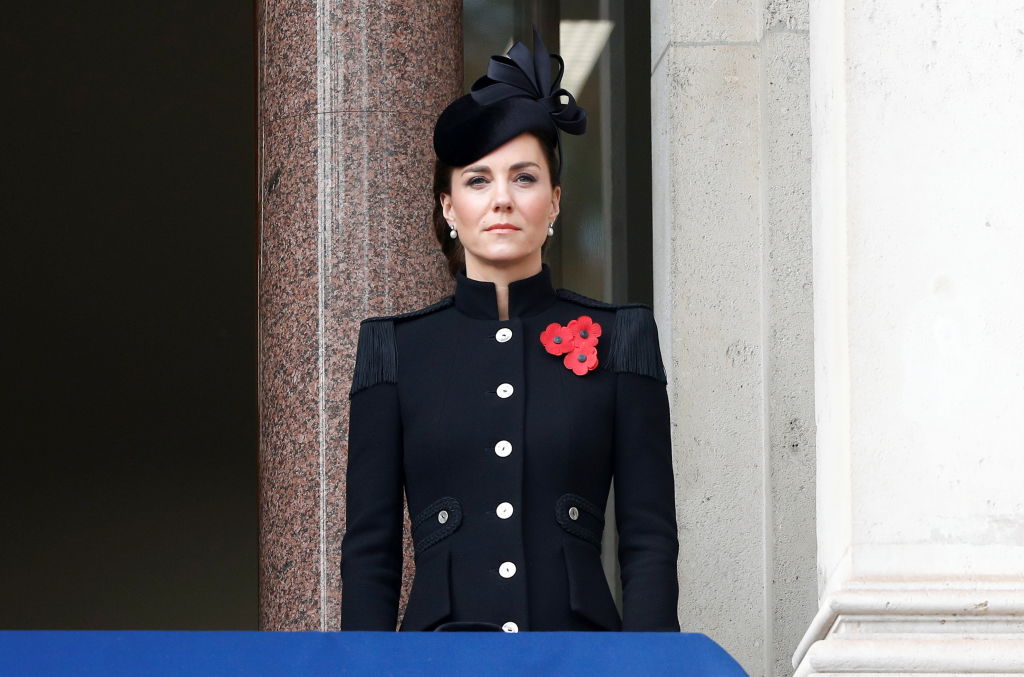 Kate Middleton îmbrăcată într-un palton negru cu guler nalt și cu flori roșii în piept și cu o pălărie neagră