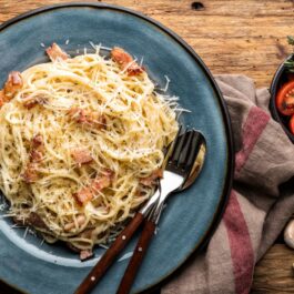 O farfurie albastră cu spaghete carbonara, pe care sunt așezate două tacâmuri. Preparatul este așezat pe o masă, alături de un prosop de bucătărie gri, câtiva căței de usturoi și un bol cu roșii tăiate și rozmarin.