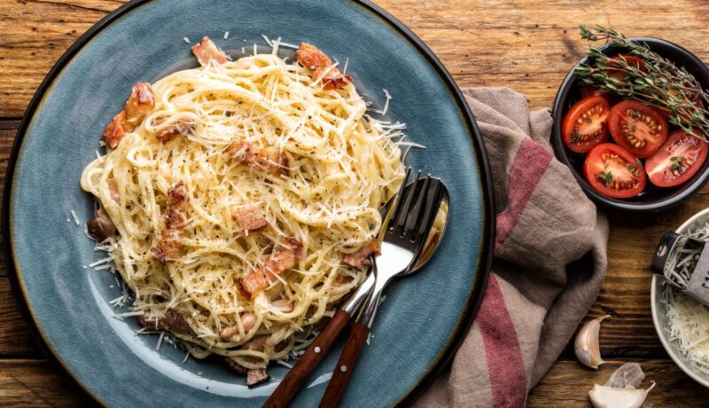 O farfurie albastră cu spaghete carbonara, pe care sunt așezate două tacâmuri. Preparatul este așezat pe o masă, alături de un prosop de bucătărie gri, câtiva căței de usturoi și un bol cu roșii tăiate și rozmarin.