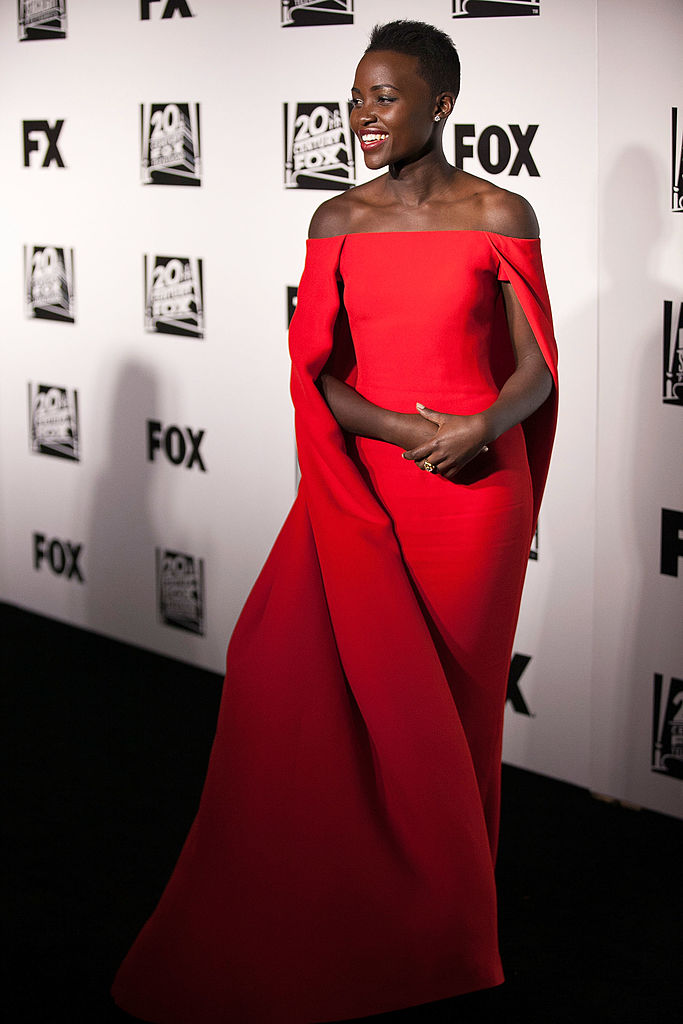 Lupita Nyongo îmbrăcată cu o rochie lungă roșie cu umerii căzuți și capă.