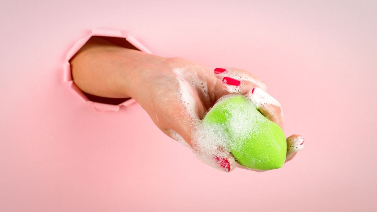 mână de femeie, cu unghii roșii, ține un burete de machiaj verde, care are pe el spună de curățare.