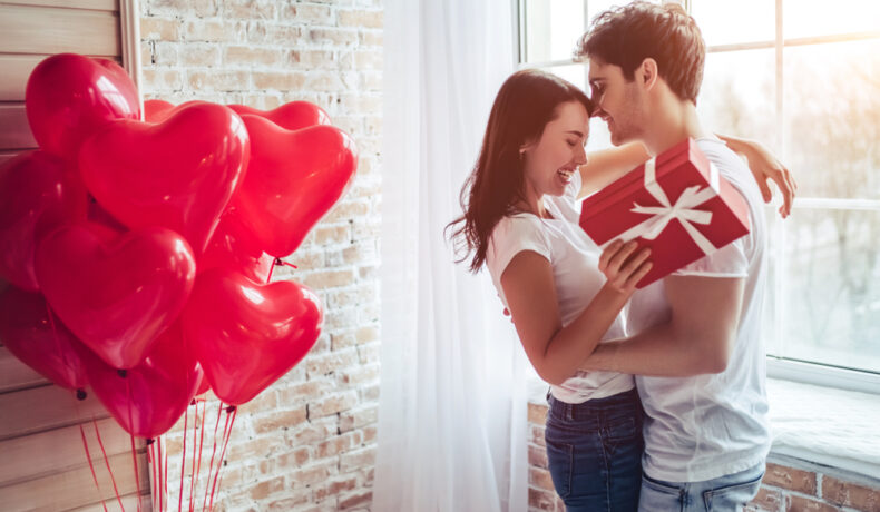 Imagine cu o cameră în care sunt baloane în care o femeie îi oferă iubitului cadoul legat de tradiții de Ziua Îndrăgostiților