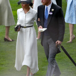 Meghan Markle îmbrăcată cu o rochie albă și o pălărie albă pe cap la brațul Prințului Harry îmbrăcat cu frac și joben